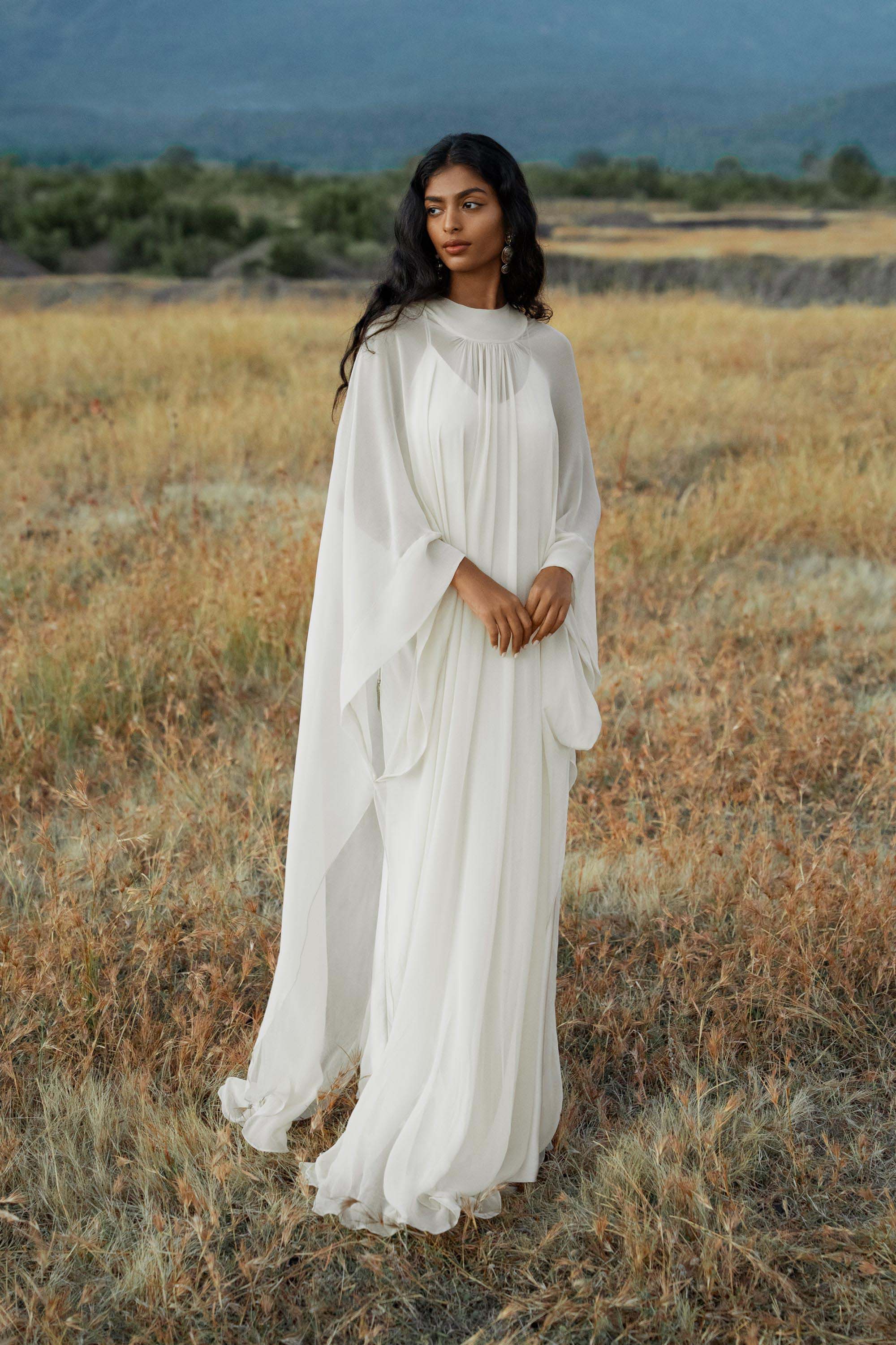 A woman with long, wavy dark hair stands in a sunlit grassy field wearing the AYA Sacred Wear Mahadevi Dress Cream—an elegant pure silk chiffon goddess gown—against a serene backdrop of hills and blue sky, gazing softly ahead with hands gently clasped.