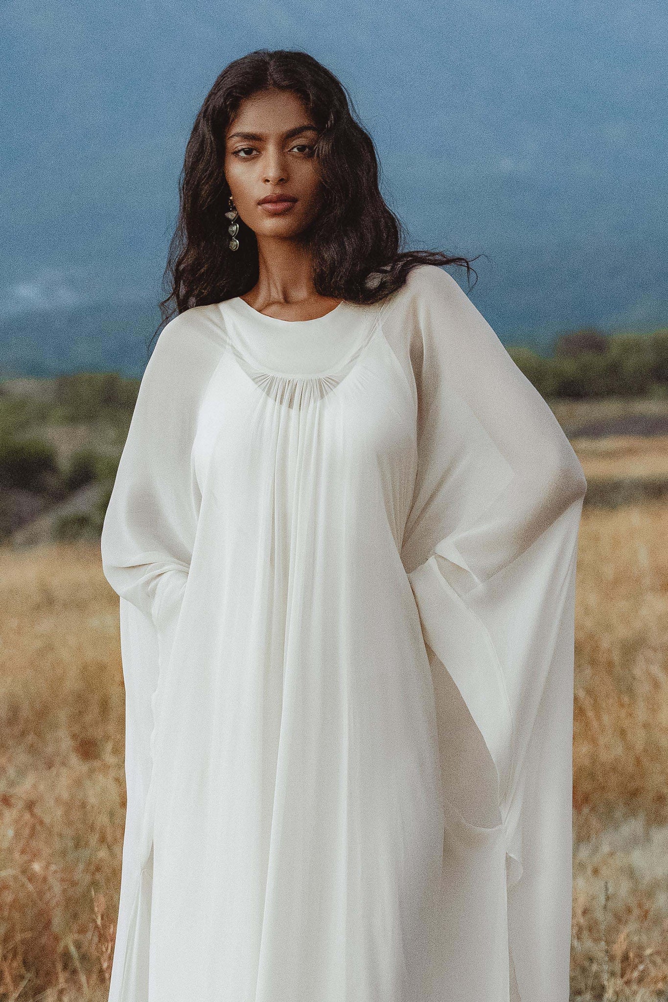 A woman with long wavy dark hair stands in a field, wearing the AYA Sacred Wear Mahadevi Dress Cream – Pure Silk Chiffon Goddess Gown. She looks serene with minimal makeup and dangling earrings, set against a blurred green-blue landscape and sky.