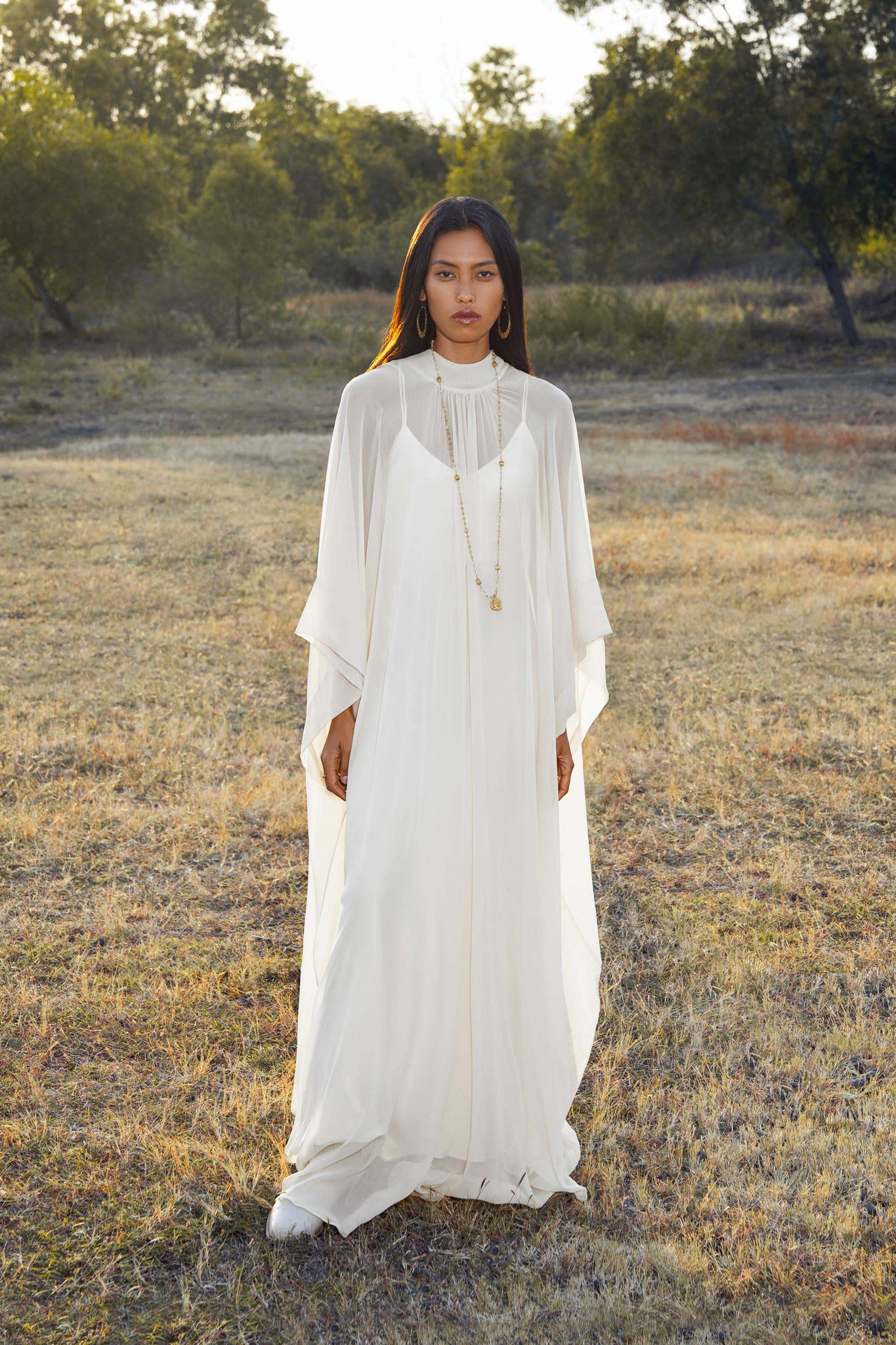A woman with long dark hair stands outdoors on dry grass, wearing the Mahadevi Dress Cream – Pure Silk Chiffon Goddess Gown by AYA Sacred Wear. She accessorizes with gold hoop earrings and layered necklaces; trees and sunlight fill the background.