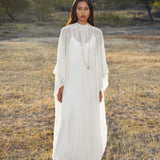 A woman with long dark hair stands outdoors on dry grass, wearing the Mahadevi Dress Cream – Pure Silk Chiffon Goddess Gown by AYA Sacred Wear. She accessorizes with gold hoop earrings and layered necklaces; trees and sunlight fill the background.