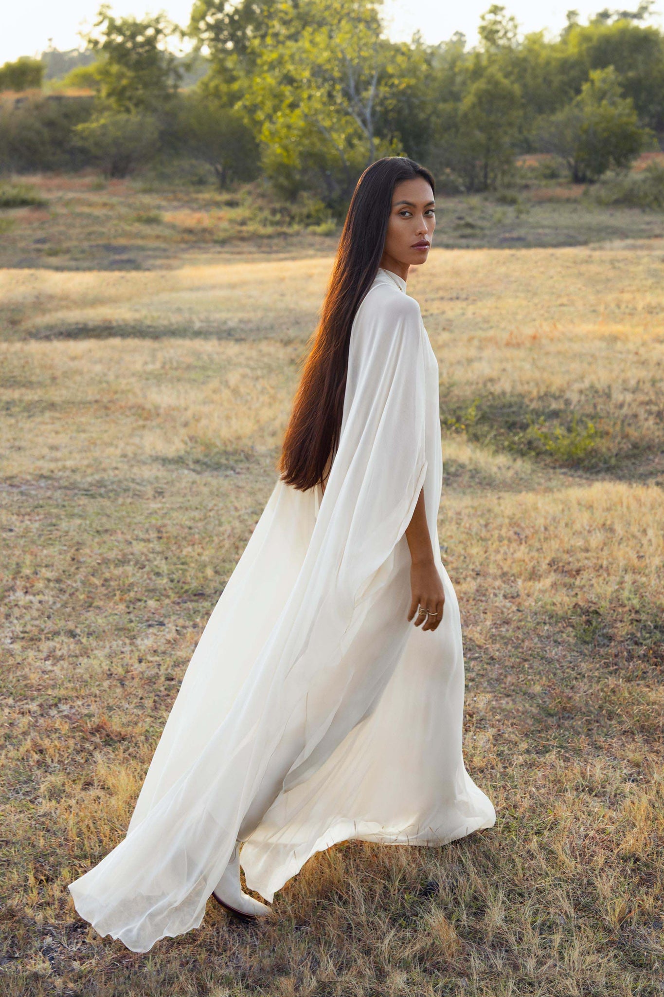 A woman with long dark hair walks through a sunlit field wearing the Mahadevi Dress Cream – Pure Silk Chiffon Goddess Gown by AYA Sacred Wear. Trees and shrubs frame her as she looks gracefully over her shoulder at the camera.