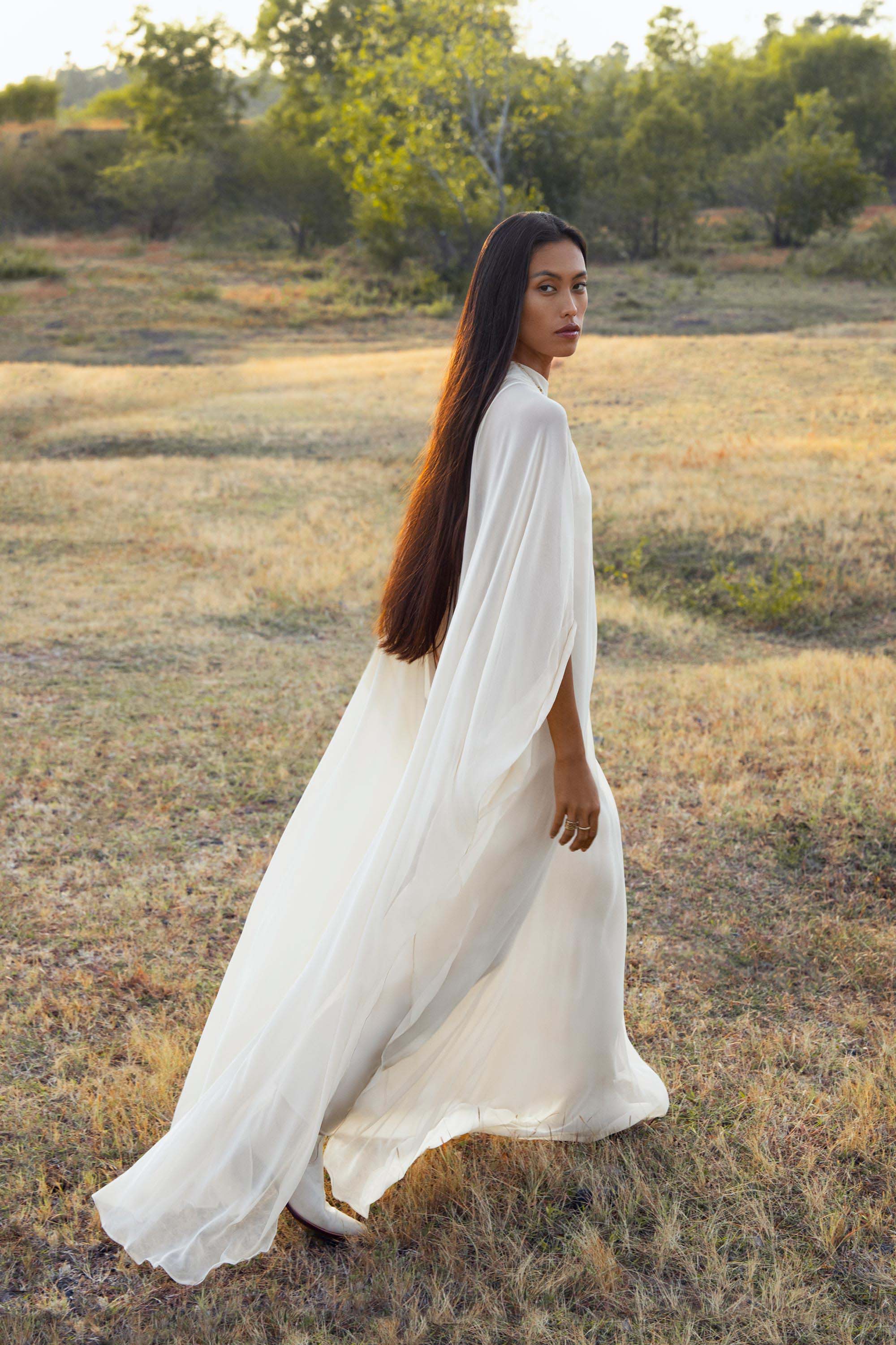 A woman with long dark hair walks through a sunlit field wearing the Mahadevi Dress Cream – Pure Silk Chiffon Goddess Gown by AYA Sacred Wear. Trees and shrubs frame her as she looks gracefully over her shoulder at the camera.