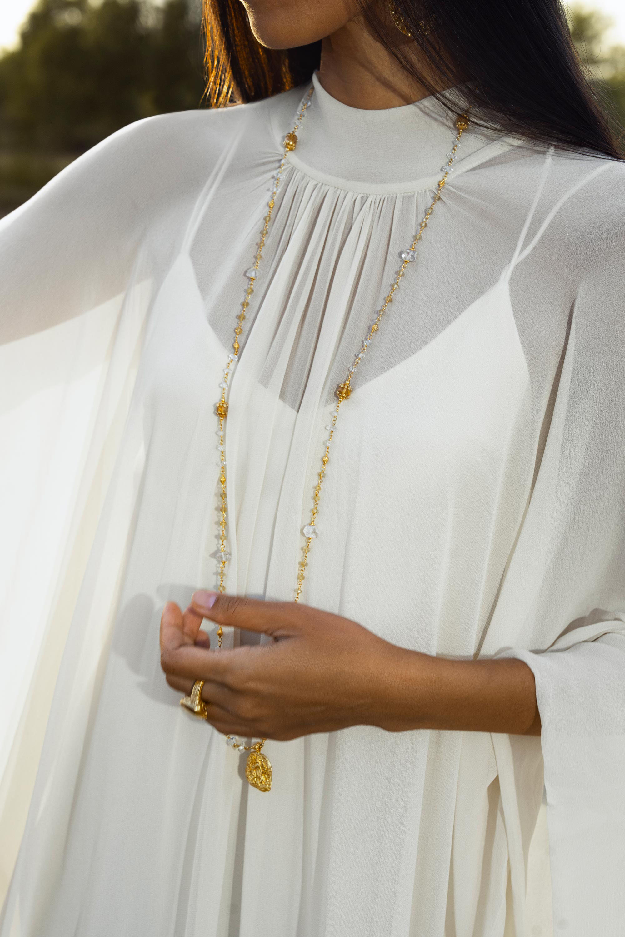 A woman stands outdoors in the Mahadevi Dress Cream – Pure Silk Chiffon Goddess Gown by AYA Sacred Wear, sunlight illuminating her sheer sleeves and delicate jewelry. Her gold and pearl necklace and rings evoke timeless luxury. Handcrafted in Bali.
