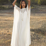 A woman stands outdoors on dry grass in the Mahadevi Dress Cream – Pure Silk Chiffon Goddess Gown by AYA Sacred Wear, raising her arms amid green trees and soft sunlight for a serene, natural scene.