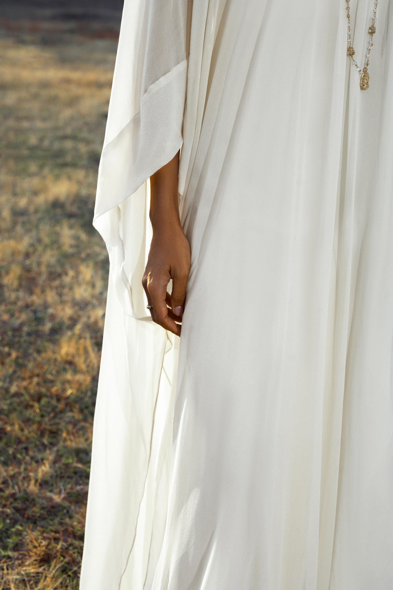 A person in the Mahadevi Dress Cream – Pure Silk Chiffon Goddess Gown by AYA Sacred Wear stands on dry grass, their hand gently touching the flowing fabric. A sheer sleeve, delicate necklace, and a softly blurred background complete the image.