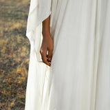 A person in the Mahadevi Dress Cream – Pure Silk Chiffon Goddess Gown by AYA Sacred Wear stands on dry grass, their hand gently touching the flowing fabric. A sheer sleeve, delicate necklace, and a softly blurred background complete the image.