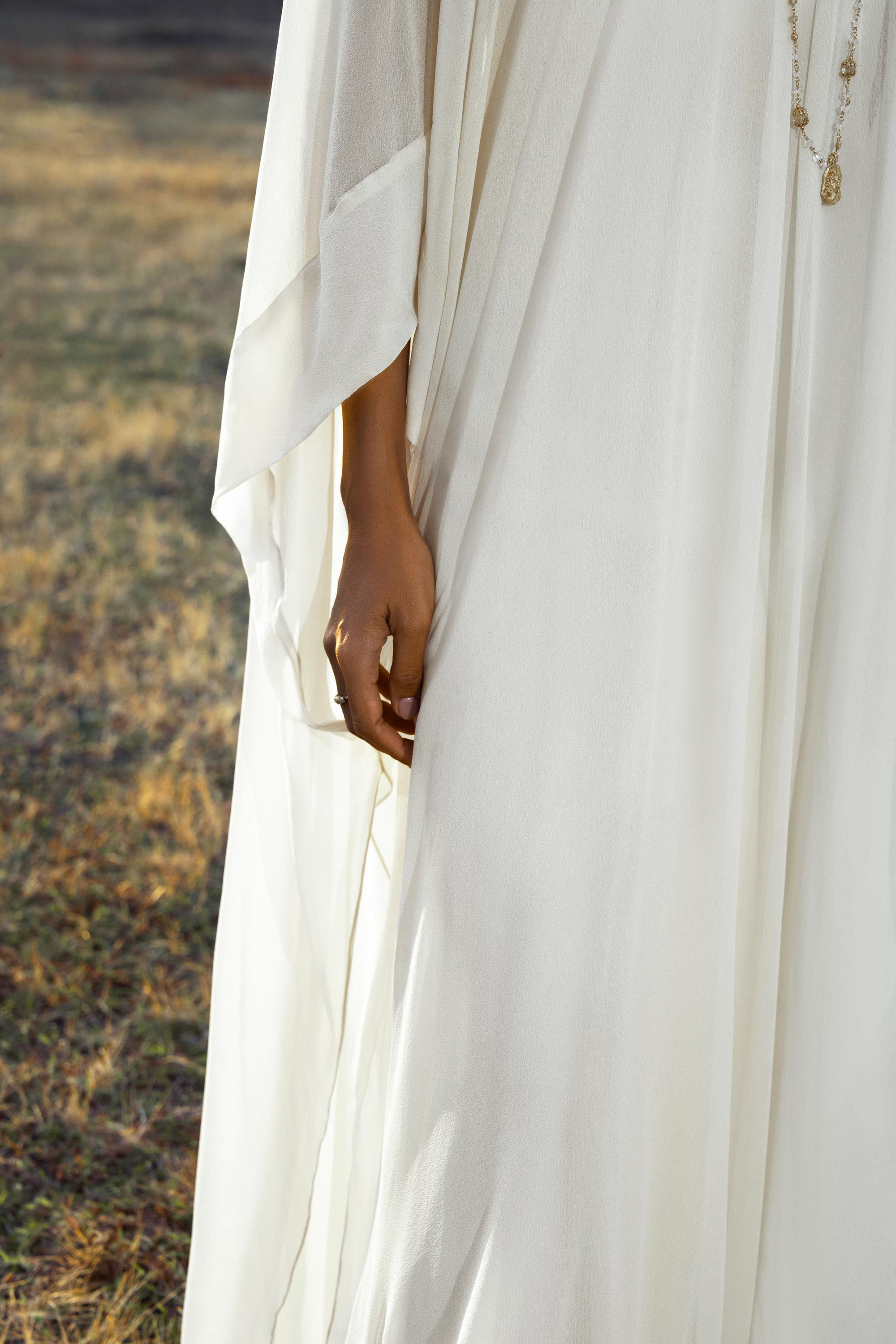 A person in the Mahadevi Dress Cream – Pure Silk Chiffon Goddess Gown by AYA Sacred Wear stands on dry grass, their hand gently touching the flowing fabric. A sheer sleeve, delicate necklace, and a softly blurred background complete the image.