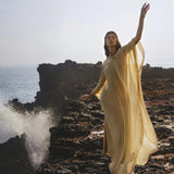 A woman in the Mahadevi Dress Inka Gold by AYA Sacred Wear, a flowing pure silk chiffon gown handcrafted in Bali, stands gracefully on seaside rocks as waves crash and mist swirls, creating a serene, ethereal scene.