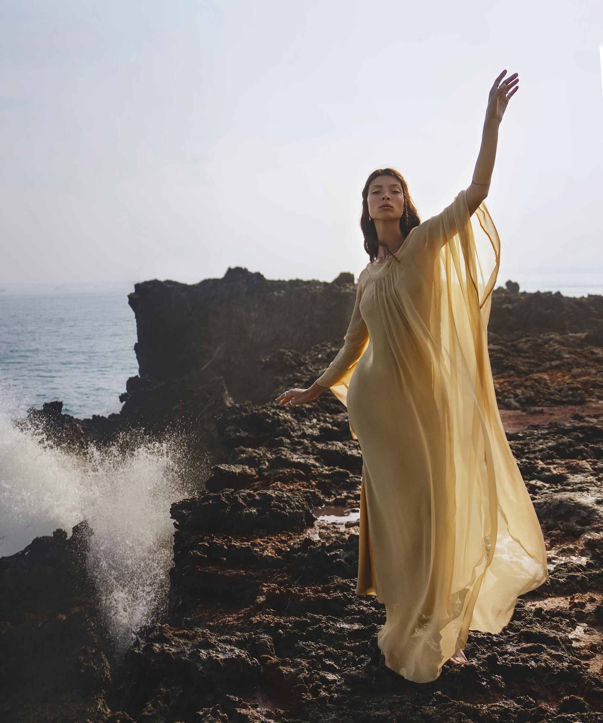 A woman in the Mahadevi Dress Inka Gold by AYA Sacred Wear, a flowing pure silk chiffon gown handcrafted in Bali, stands gracefully on seaside rocks as waves crash and mist swirls, creating a serene, ethereal scene.