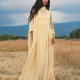 A woman with long, wavy black hair stands in a grassy field wearing the AYA Sacred Wear Mahadevi Dress Inka Gold, a pure silk chiffon goddess gown, gazing confidently at the camera with mountains and clouds in the background.