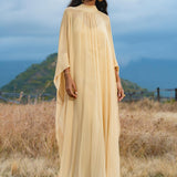 A woman stands outdoors on dry grass, wearing the Mahadevi Dress Inka Gold – a pure silk chiffon goddess gown by AYA Sacred Wear. She has long dark hair, gold hoop earrings, and is framed by a cloudy sky with a distant green mountain.