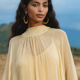 A woman with long wavy dark hair wears the AYA Sacred Wear Mahadevi Dress Inka Gold – a pure silk chiffon goddess gown, standing outdoors before blurred hills and a cloudy sky, gazing calmly at the camera.