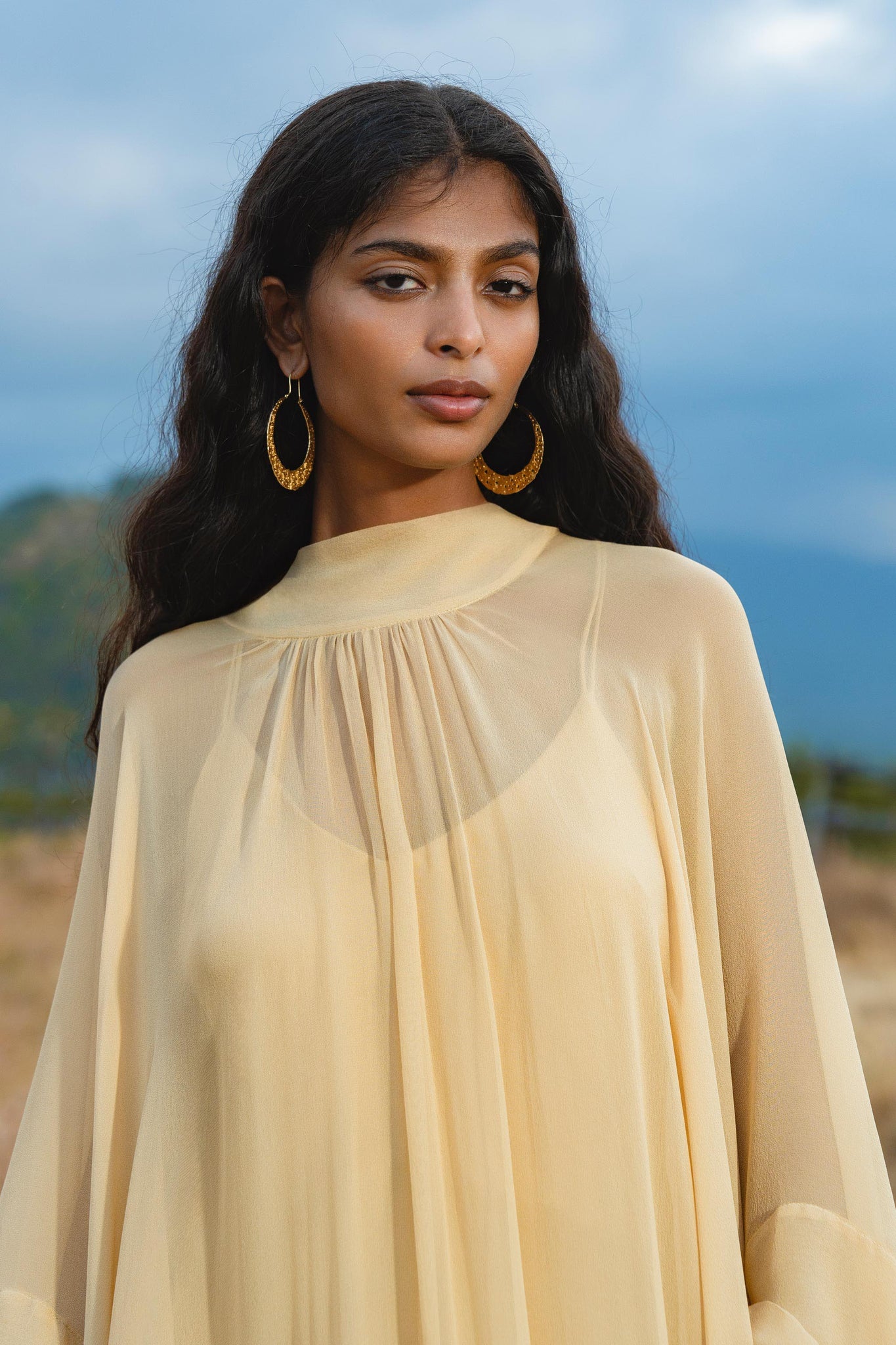 A woman with long wavy dark hair wears the AYA Sacred Wear Mahadevi Dress Inka Gold – a pure silk chiffon goddess gown, standing outdoors before blurred hills and a cloudy sky, gazing calmly at the camera.