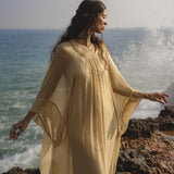 A woman in the Mahadevi Dress Inka Gold by AYA Sacred Wear, a pure silk chiffon goddess gown, stands on rocky seaside terrain with waves crashing and sunlight catching her hair, embodying serenity and ethereal elegance.