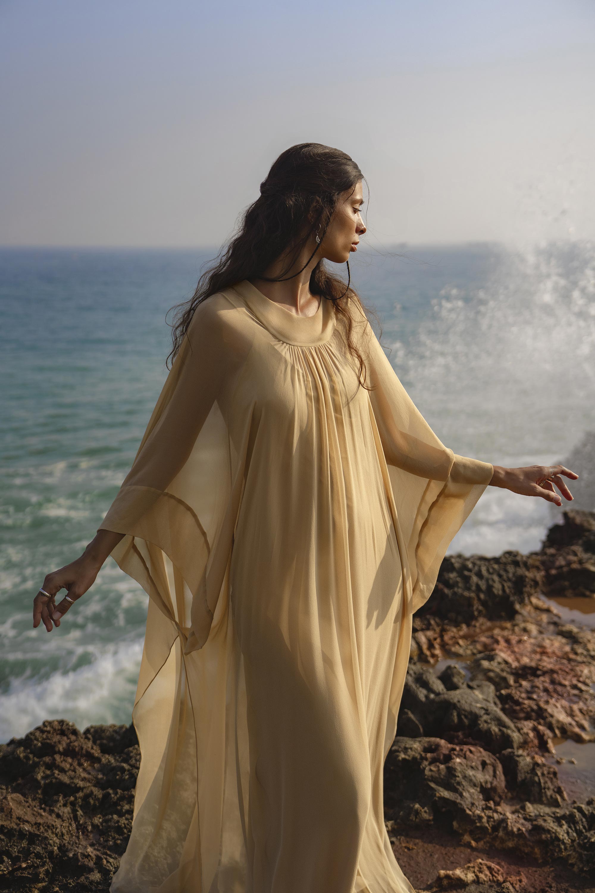 A woman in the Mahadevi Dress Inka Gold by AYA Sacred Wear, a pure silk chiffon goddess gown, stands on rocky seaside terrain with waves crashing and sunlight catching her hair, embodying serenity and ethereal elegance.