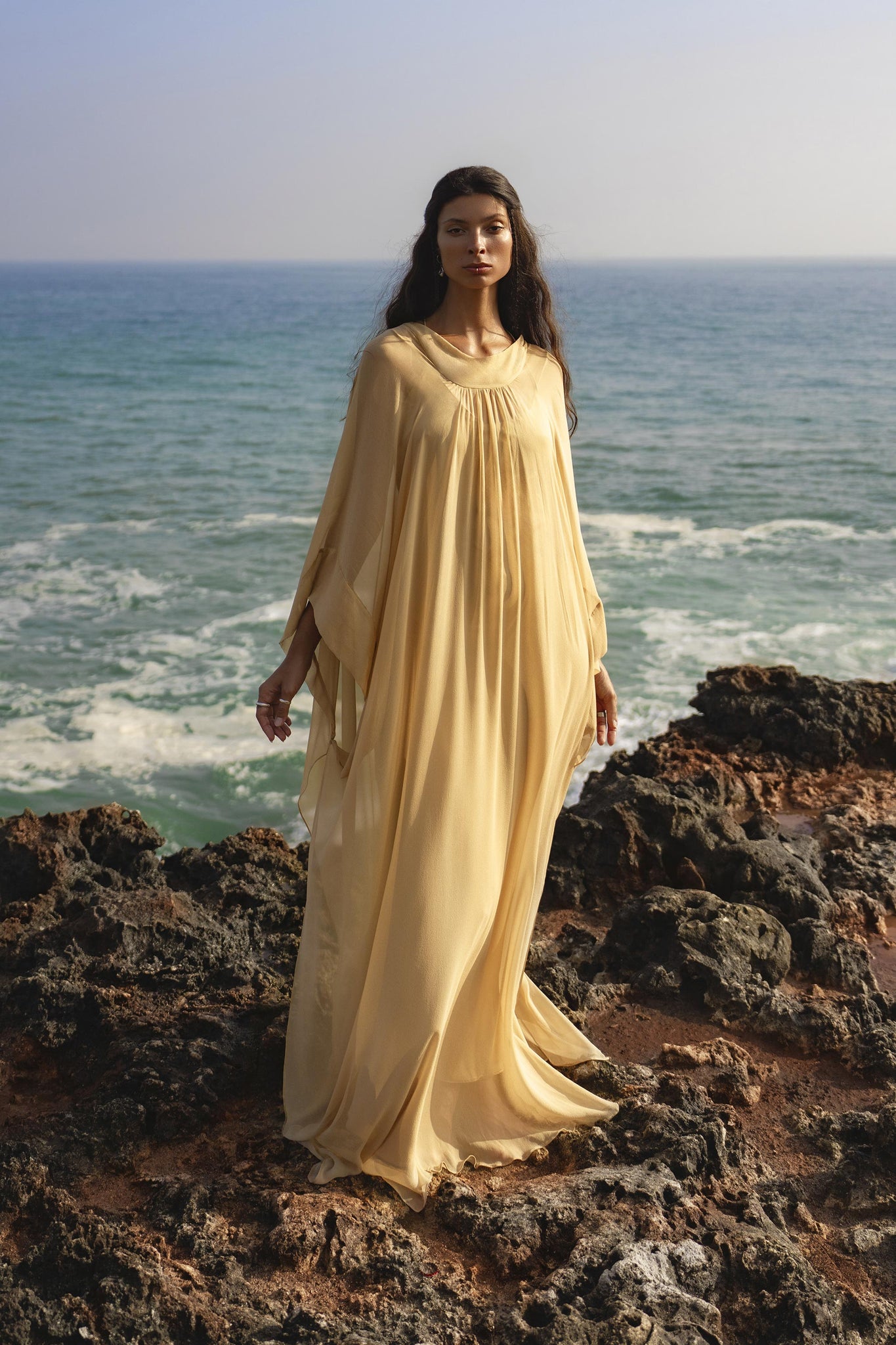 A woman with long, dark hair stands on rocky shorelines in a flowing AYA Sacred Wear Mahadevi Dress Inka Gold, a pure silk chiffon goddess gown. Ocean waves crash behind her as her dress billows gracefully in the breeze beneath clear skies.