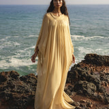 A woman with long, dark hair stands on rocky shorelines in a flowing AYA Sacred Wear Mahadevi Dress Inka Gold, a pure silk chiffon goddess gown. Ocean waves crash behind her as her dress billows gracefully in the breeze beneath clear skies.