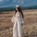 A woman wearing the Maharani Blouse in Cream Colour – Pure Silk Chiffon Boho Top by AYA Sacred Wear stands in a grassy field, cream feathered hat on her curly hair, holding a book near cloud-capped mountains under a blue sky.