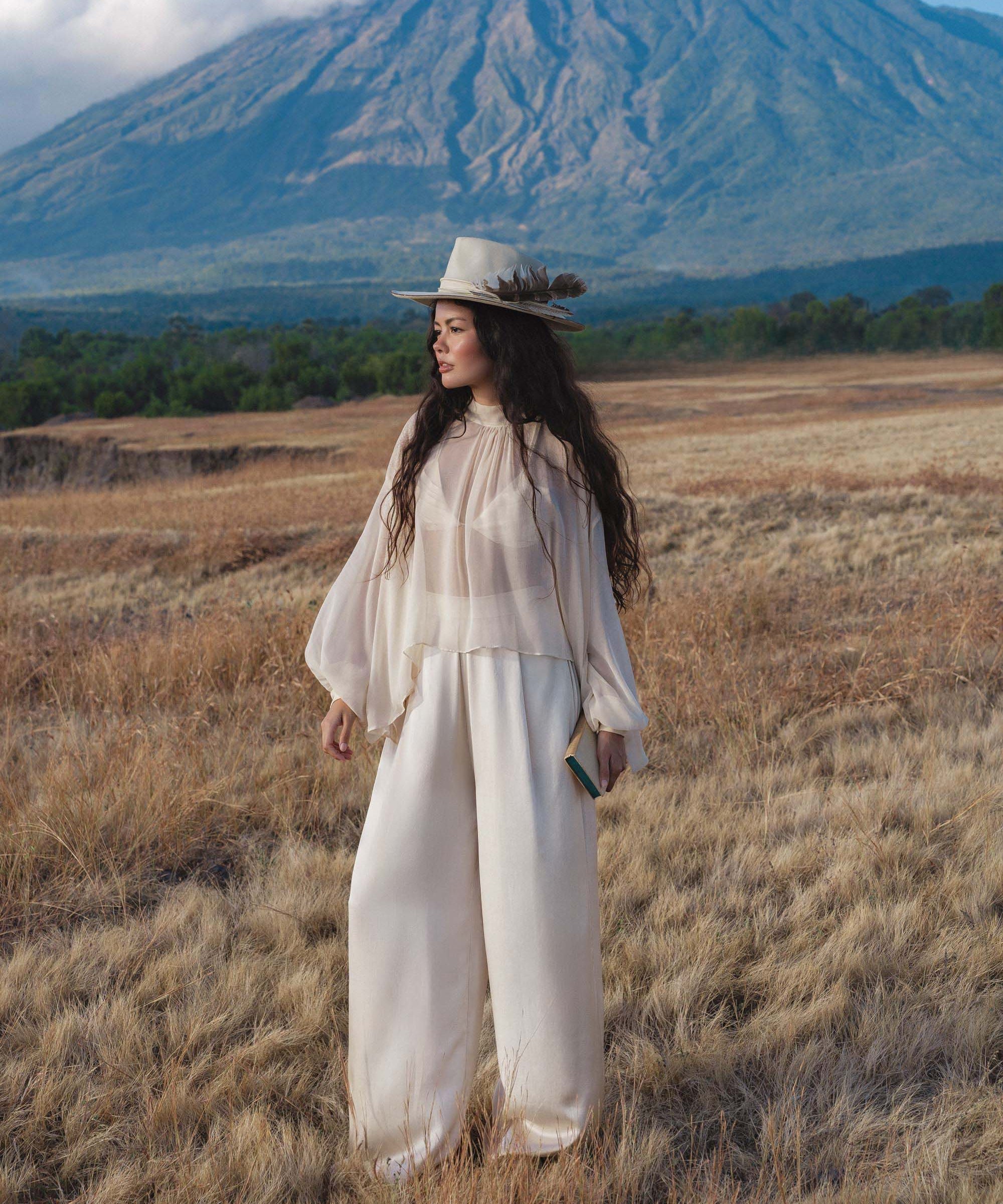A woman wearing the Maharani Blouse in Cream Colour – Pure Silk Chiffon Boho Top by AYA Sacred Wear stands in a grassy field, cream feathered hat on her curly hair, holding a book near cloud-capped mountains under a blue sky.