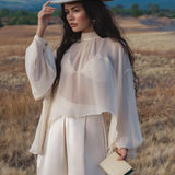 A woman in the AYA Sacred Wear Maharani Blouse in Cream – a pure silk chiffon boho top – and wide-leg silk pants stands in a dry field, wearing a feathered cream hat, holding a beige clutch, with mountains under a blue sky behind her.