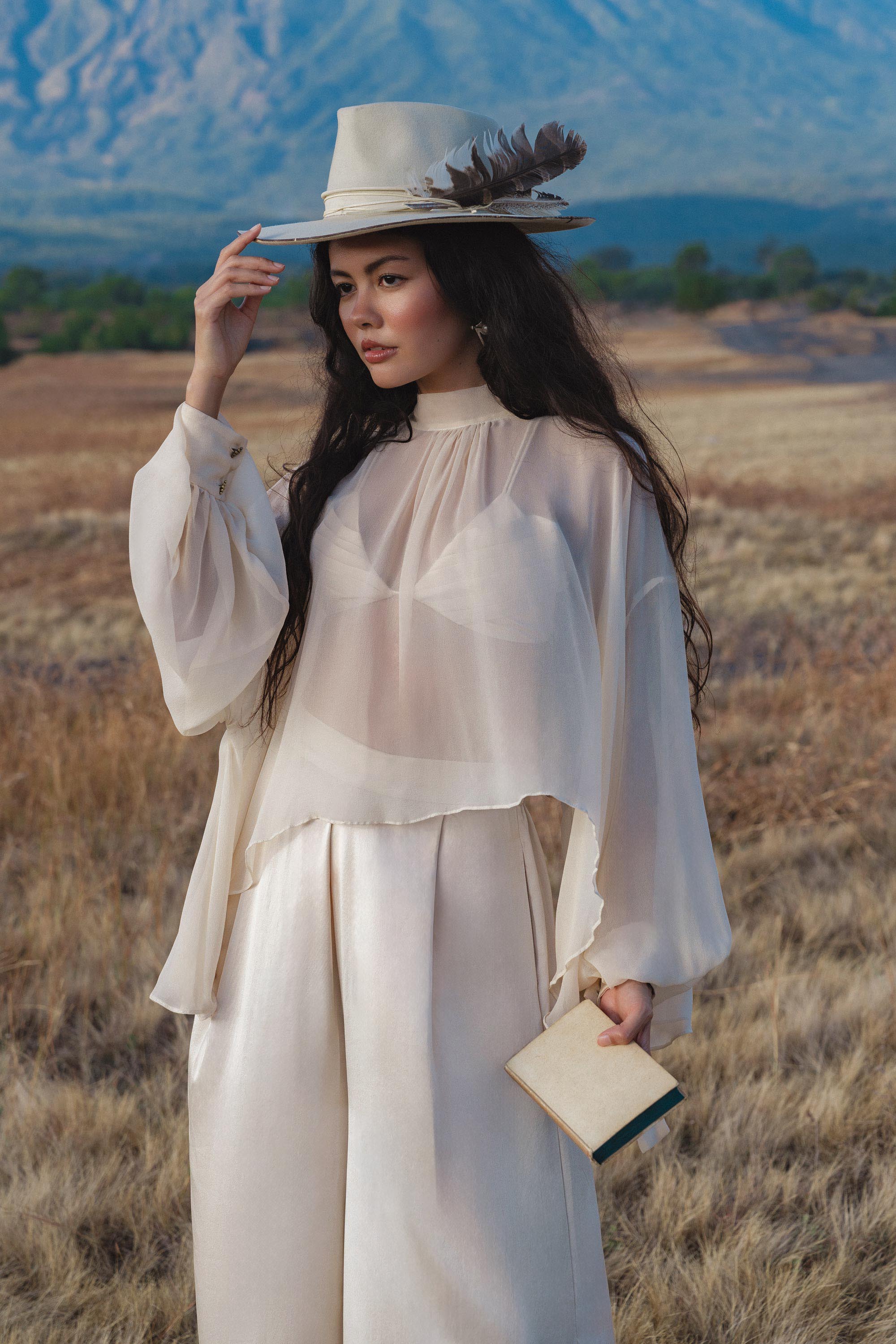 A woman in the AYA Sacred Wear Maharani Blouse in Cream – a pure silk chiffon boho top – and wide-leg silk pants stands in a dry field, wearing a feathered cream hat, holding a beige clutch, with mountains under a blue sky behind her.