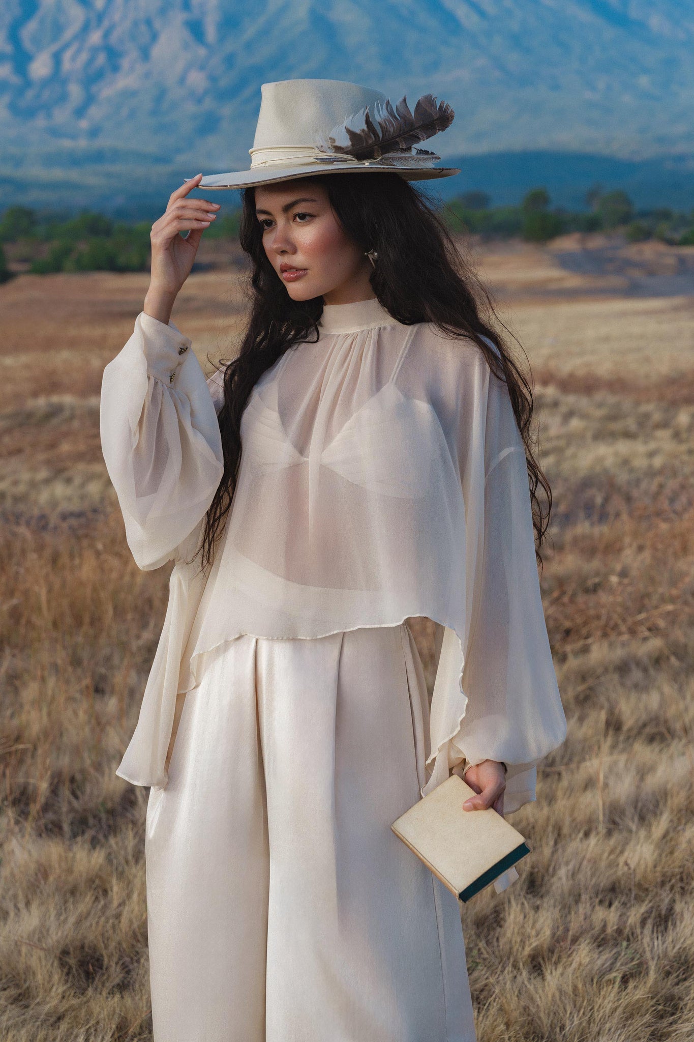 A woman in the AYA Sacred Wear Maharani Blouse in Cream – a pure silk chiffon boho top – and wide-leg silk pants stands in a dry field, wearing a feathered cream hat, holding a beige clutch, with mountains under a blue sky behind her.