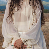 A woman with long, wavy dark hair wears the Maharani Blouse in Cream Colour – Pure Silk Chiffon Boho Top by AYA Sacred Wear. She stands outdoors in a field, holding an open book with mountains softly blurred behind her.
