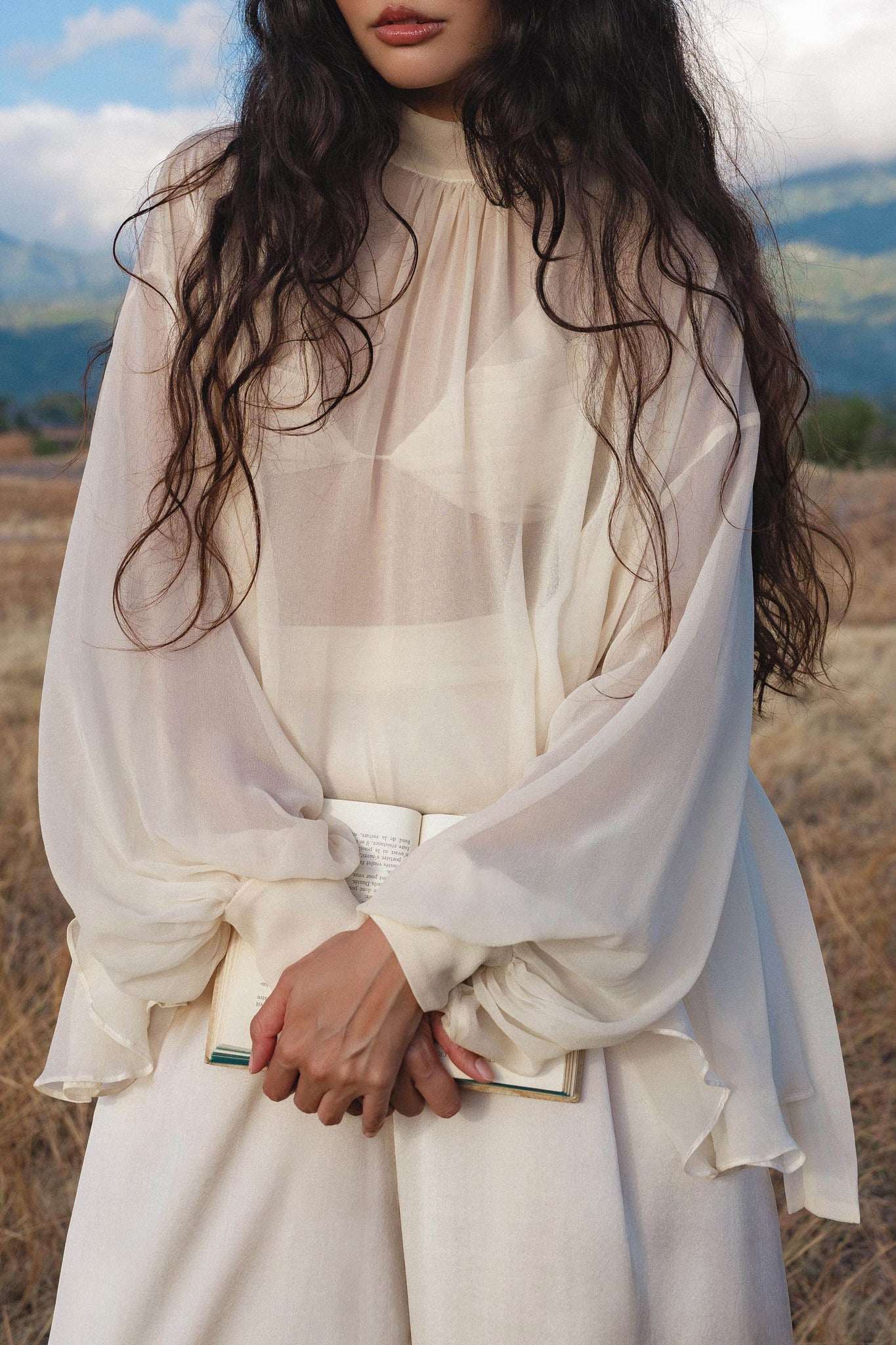 A woman with long, wavy dark hair wears the Maharani Blouse in Cream Colour – Pure Silk Chiffon Boho Top by AYA Sacred Wear. She stands outdoors in a field, holding an open book with mountains softly blurred behind her.