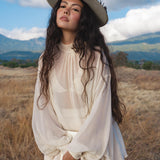 A young woman with long, wavy dark hair stands in a grassy field before mountains and clouds, wearing a wide-brimmed hat and the Maharani Blouse in Cream Colour—Pure Silk Chiffon Boho Top by AYA Sacred Wear, looking confidently at the camera.