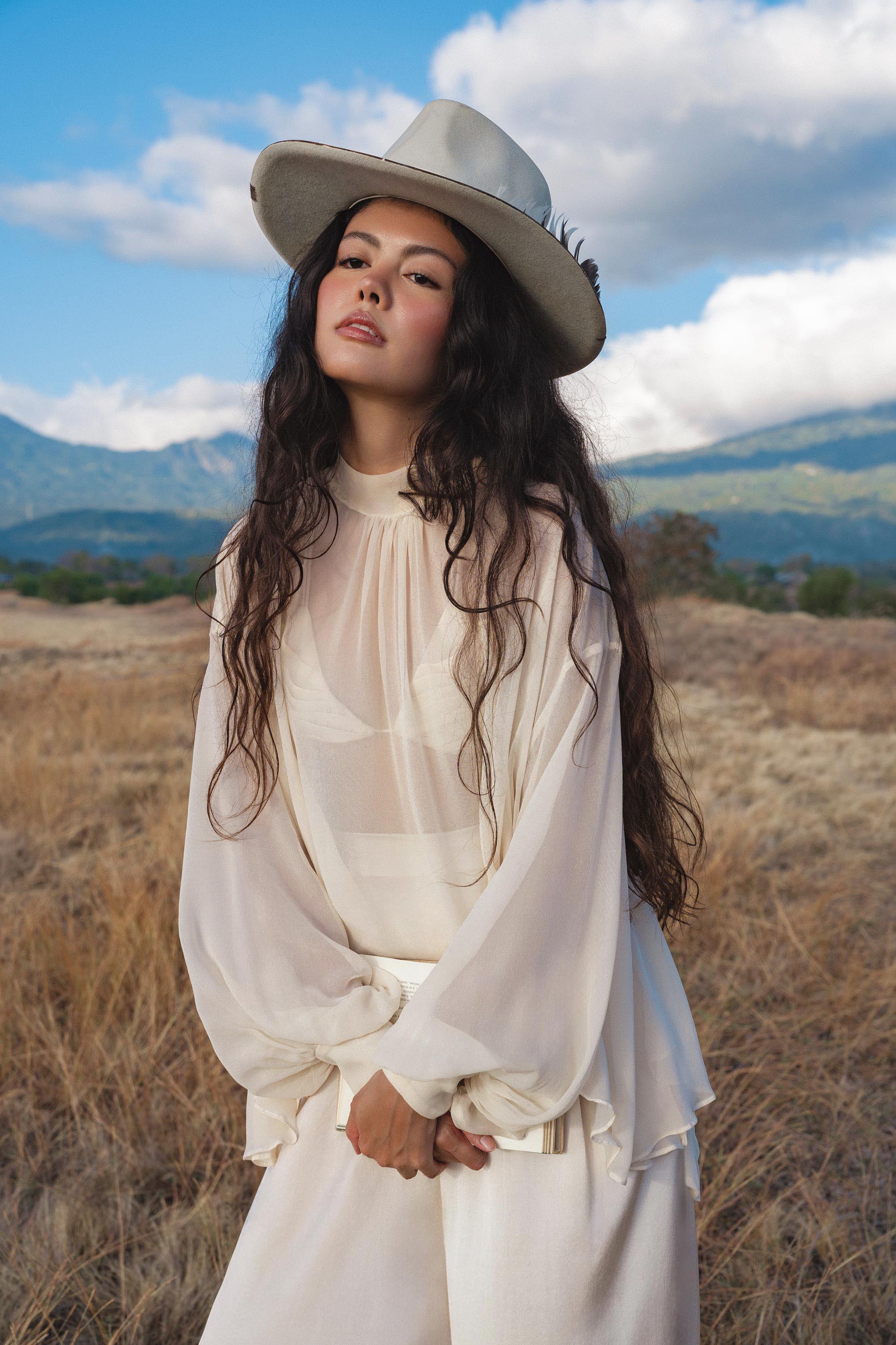 A young woman with long, wavy dark hair stands in a grassy field before mountains and clouds, wearing a wide-brimmed hat and the Maharani Blouse in Cream Colour—Pure Silk Chiffon Boho Top by AYA Sacred Wear, looking confidently at the camera.