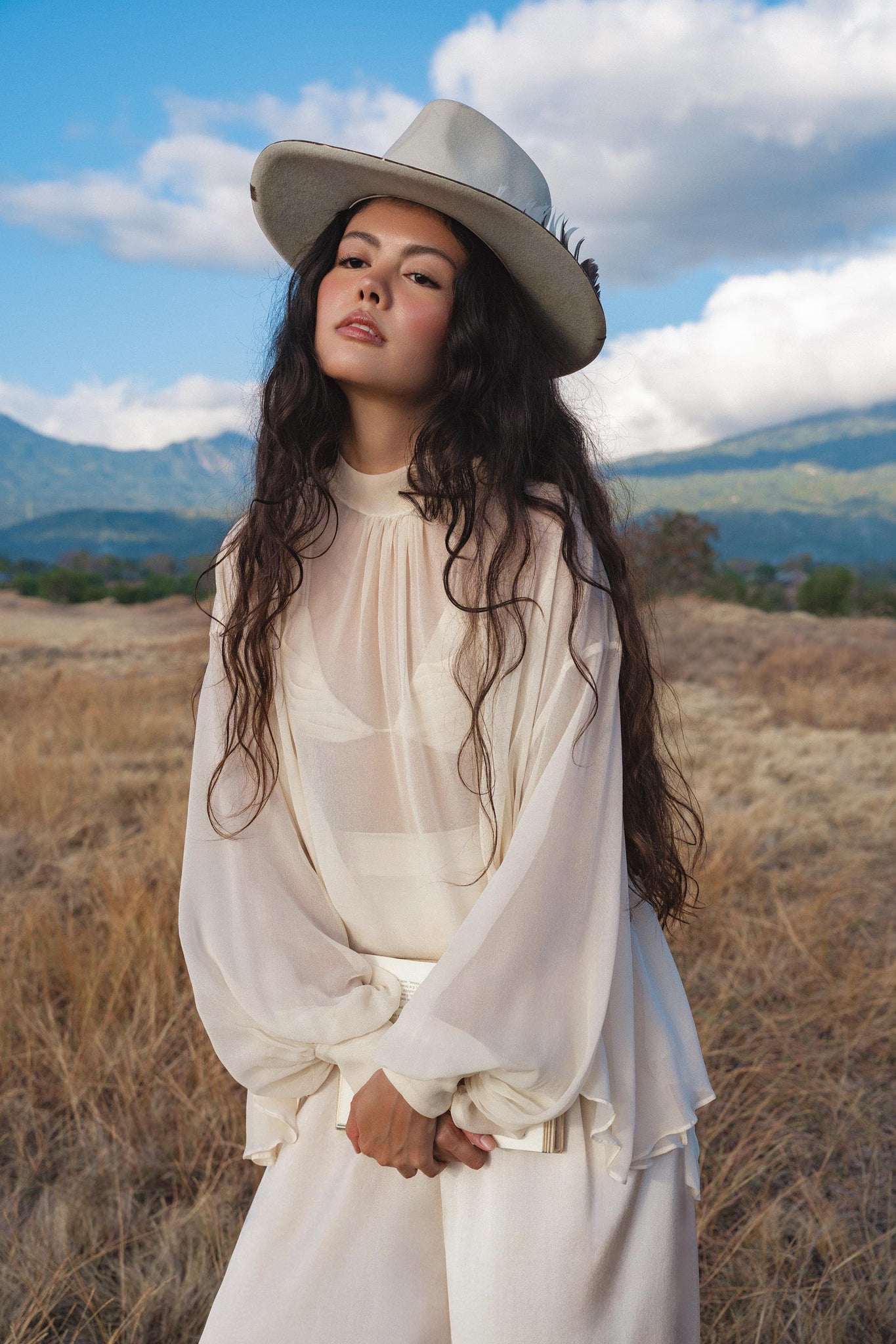 A young woman with long, wavy dark hair stands in a grassy field before mountains and clouds, wearing a wide-brimmed hat and the Maharani Blouse in Cream Colour—Pure Silk Chiffon Boho Top by AYA Sacred Wear, looking confidently at the camera.