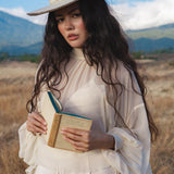 A woman with long wavy hair wears the Maharani Blouse in Cream Colour – Pure Silk Chiffon Boho Top by AYA Sacred Wear and a feathered wide-brim hat. She stands in a dry grassy field, holding an open book before a backdrop of mountains and cloudy sky.