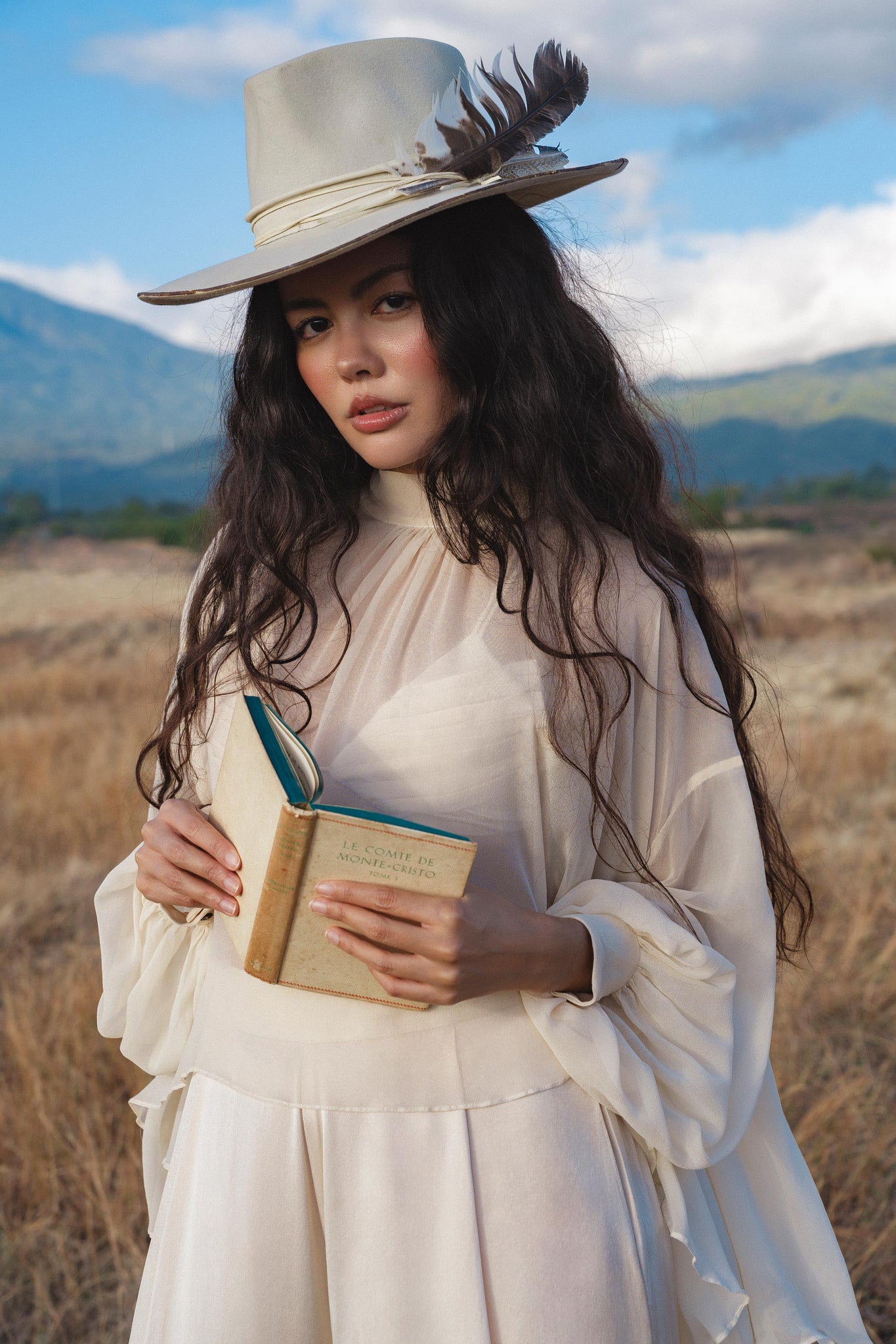 A woman with long wavy hair wears the Maharani Blouse in Cream Colour – Pure Silk Chiffon Boho Top by AYA Sacred Wear and a feathered wide-brim hat. She stands in a dry grassy field, holding an open book before a backdrop of mountains and cloudy sky.
