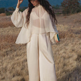In a grassy field, a woman with long wavy hair wears the Maharani Blouse in Cream Colour – Pure Silk Chiffon Boho Top by AYA Sacred Wear, wide pants, and a feathered hat. She holds a teal book with mountains and clouds as her backdrop.
