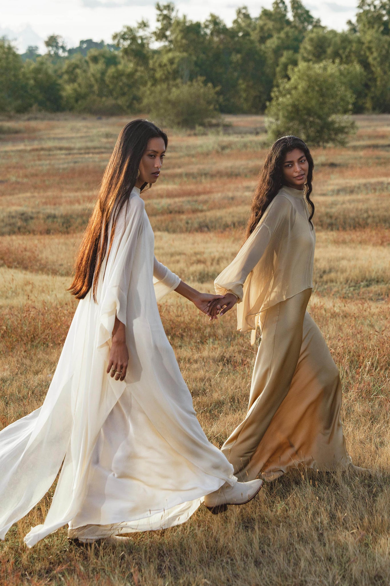 Two women walk hand in hand through a sunlit grassy field, wearing AYA Sacred Wear’s Maharani Blouse in Inka Gold—pure silk chiffon tops that flow gracefully amid the serene natural beauty.