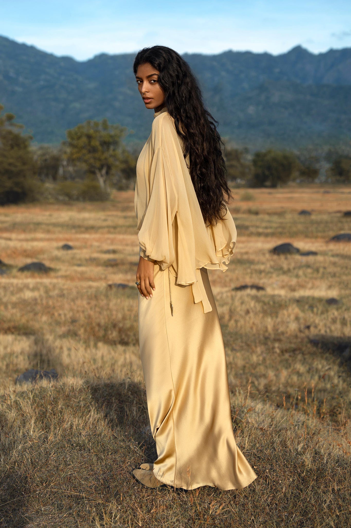 A woman with long, wavy dark hair stands in a sunlit field wearing the Maharani Blouse in Inka Gold— a pure silk chiffon boho top by AYA Sacred Wear. She gazes over her shoulder, with mountains and trees under a bright blue sky behind her.