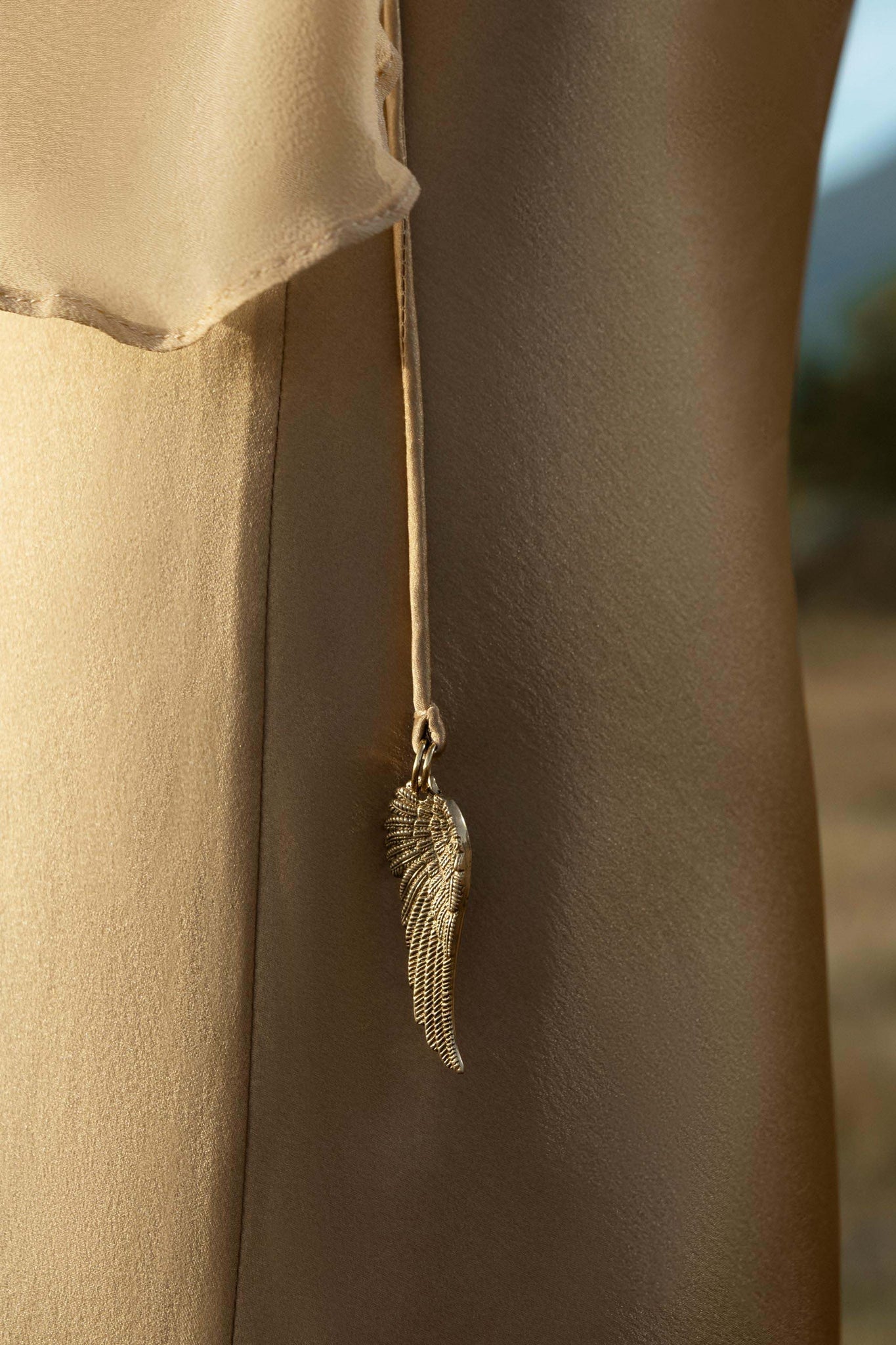 A close-up of the AYA Sacred Wear Maharani Blouse in Inka Gold—crafted from pure silk chiffon—shows a delicate gold angel wing pendant on a cord, with soft natural light enhancing the sheen against a blurred outdoor background.