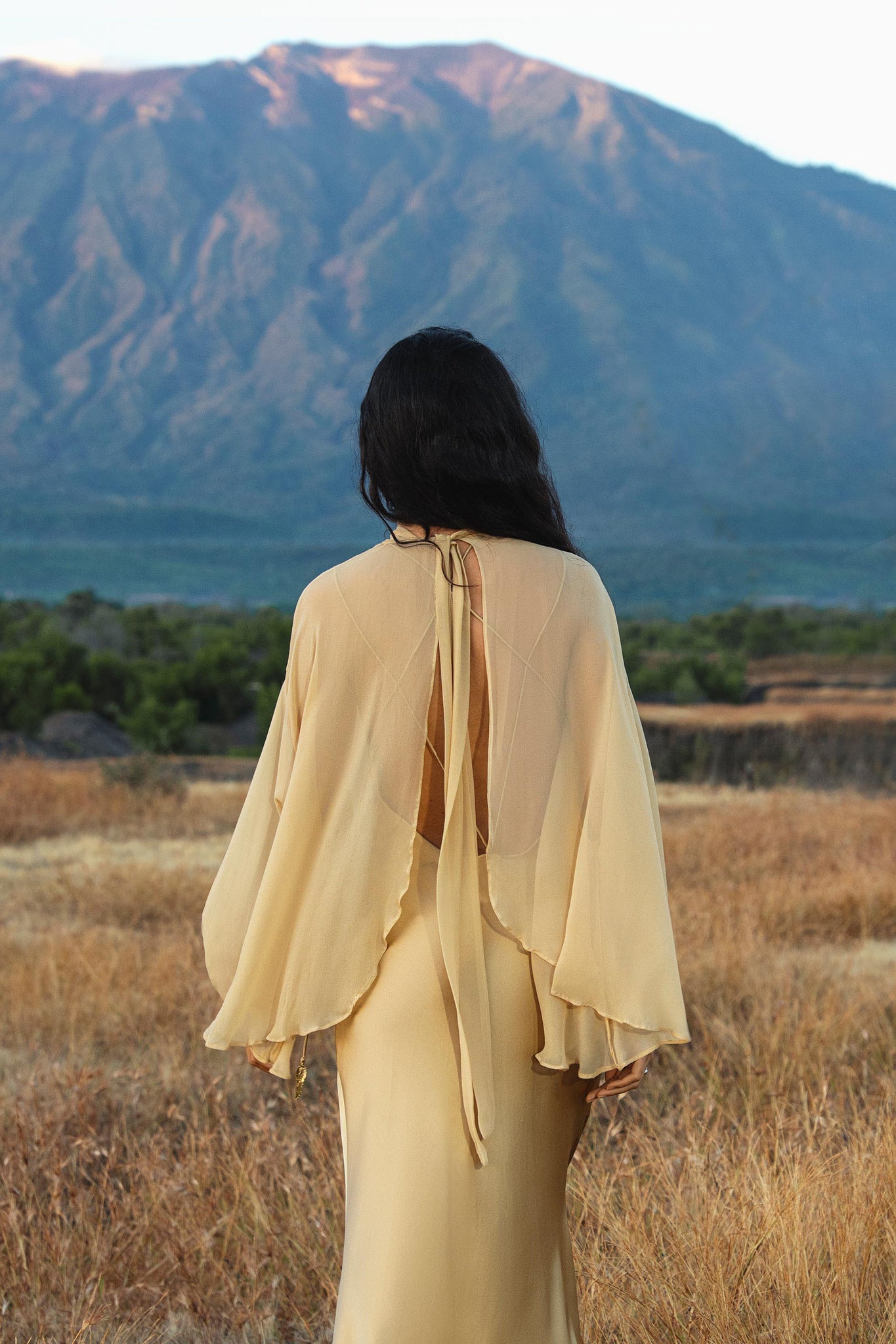 A person with long, dark hair stands in a dry grassy field, wearing the AYA Sacred Wear Maharani Blouse in Inka Gold—a pure silk chiffon boho top with loose sleeves. Behind them, a sunlit mountain rises under a clear sky.