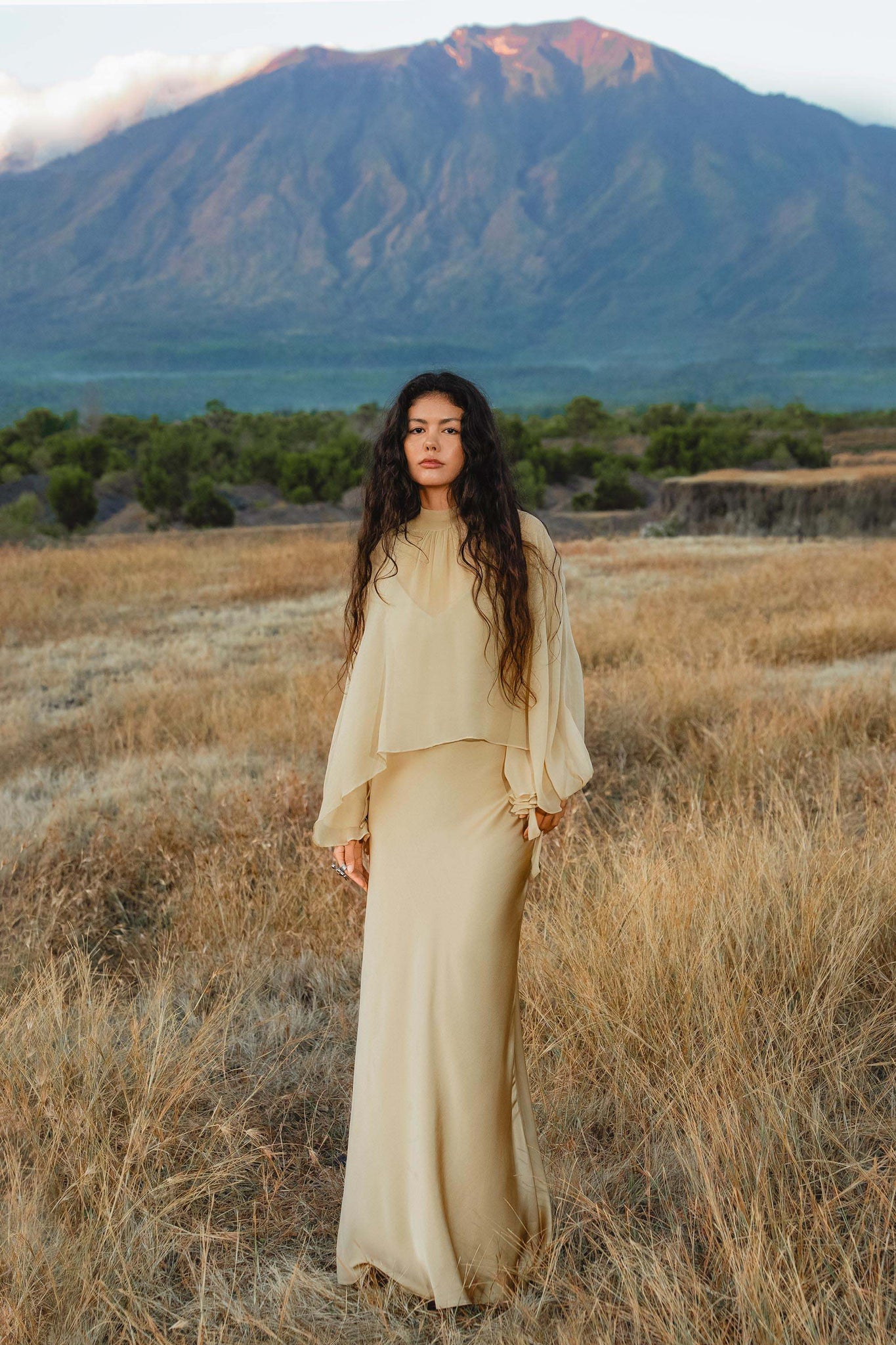 A woman with long dark curly hair stands in a dry field, wearing the flowing Maharani Blouse in Inka Gold by AYA Sacred Wear, handcrafted from pure silk chiffon. She faces the camera as green trees and misty mountains rise under a cloudy sky.