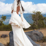 A woman with long dark hair stands on dry grass and rocks, wearing the Mandara Dress in Ivory Colour – Organic Boho Wedding Dress by AYA Sacred Wear, featuring hand-stitched embroidery, fringe, and open back details amid hills and a partly cloudy sky.