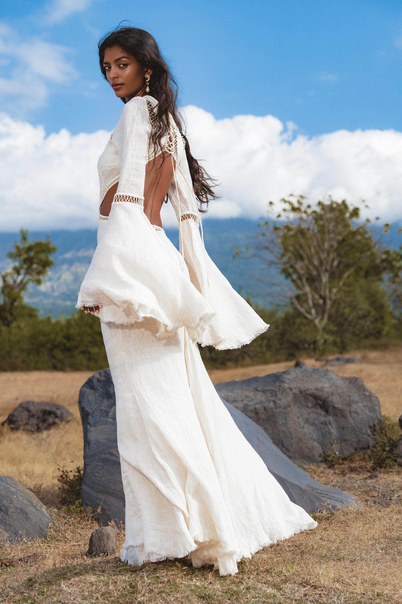 A woman with long dark hair stands on dry grass and rocks, wearing the Mandara Dress in Ivory Colour – Organic Boho Wedding Dress by AYA Sacred Wear, featuring hand-stitched embroidery, fringe, and open back details amid hills and a partly cloudy sky.