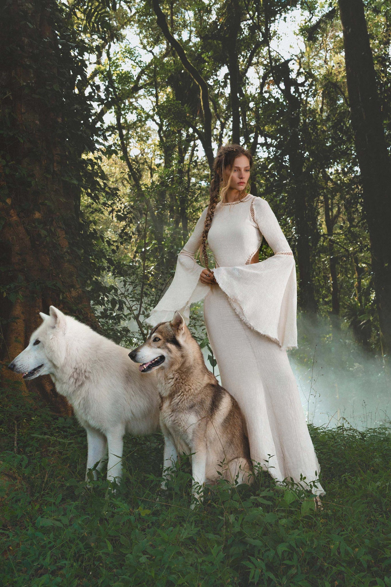 A woman in an ivory Mandara Dress by AYA Sacred Wear, featuring hand-stitched embroidery, stands in a misty green forest with two large wolves. Sunlight streams through tall trees, casting a mystical boho wedding scene.