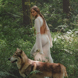 A woman wearing the Mandara Dress in Ivory Colour by AYA Sacred Wear—an organic boho wedding dress with hand-stitched embroidery—stands in a green forest, her braided hair adorned with flowers, a wolf-like dog by her side.