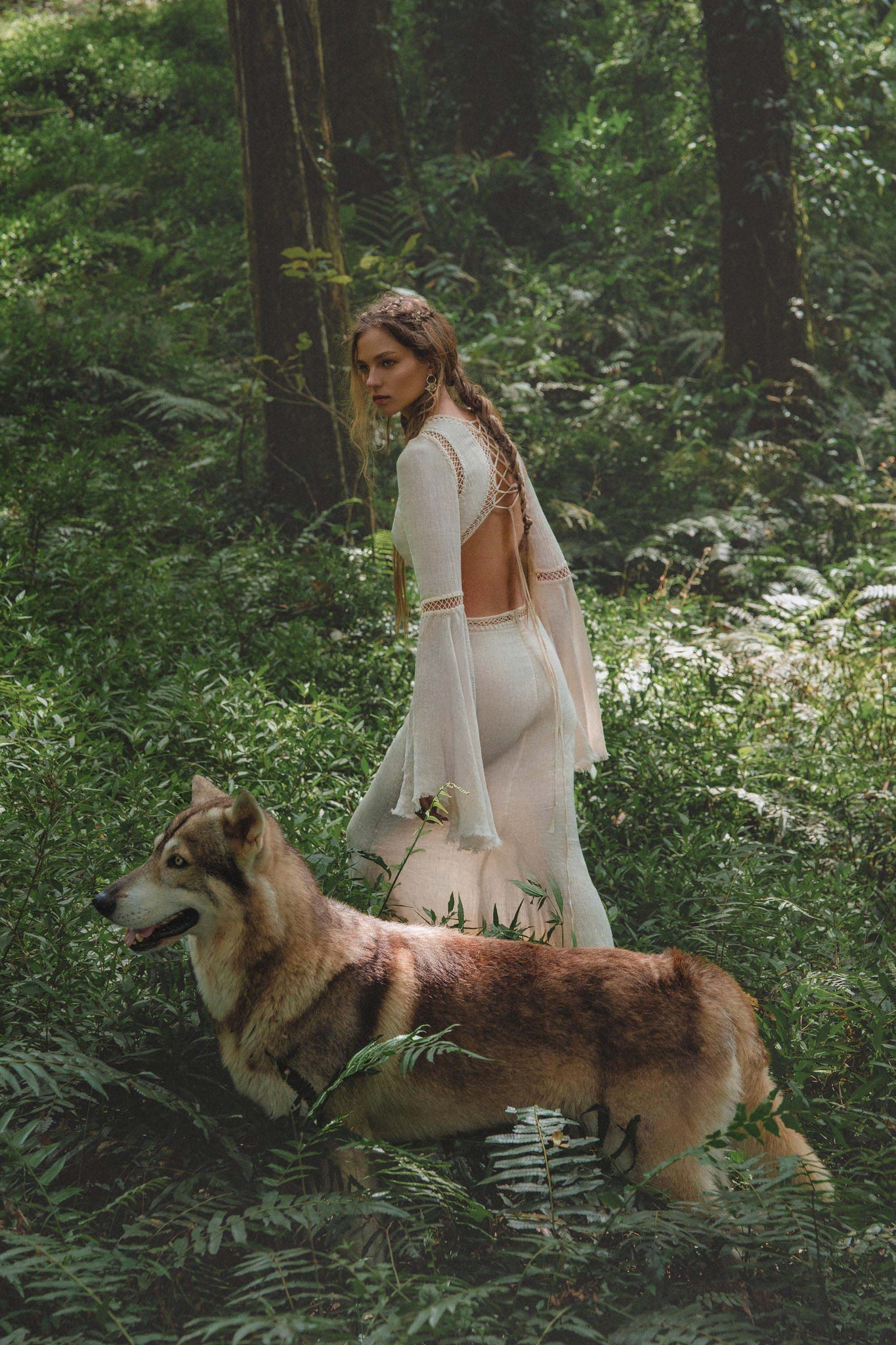 A woman wearing the Mandara Dress in Ivory Colour by AYA Sacred Wear—an organic boho wedding dress with hand-stitched embroidery—stands in a green forest, her braided hair adorned with flowers, a wolf-like dog by her side.