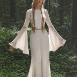 A woman with long, braided hair stands in a sunlit forest, eyes closed in serenity. She wears the AYA Sacred Wear Mandara Dress in Ivory – an organic boho wedding dress with wide bell sleeves and hand-stitched embroidery.