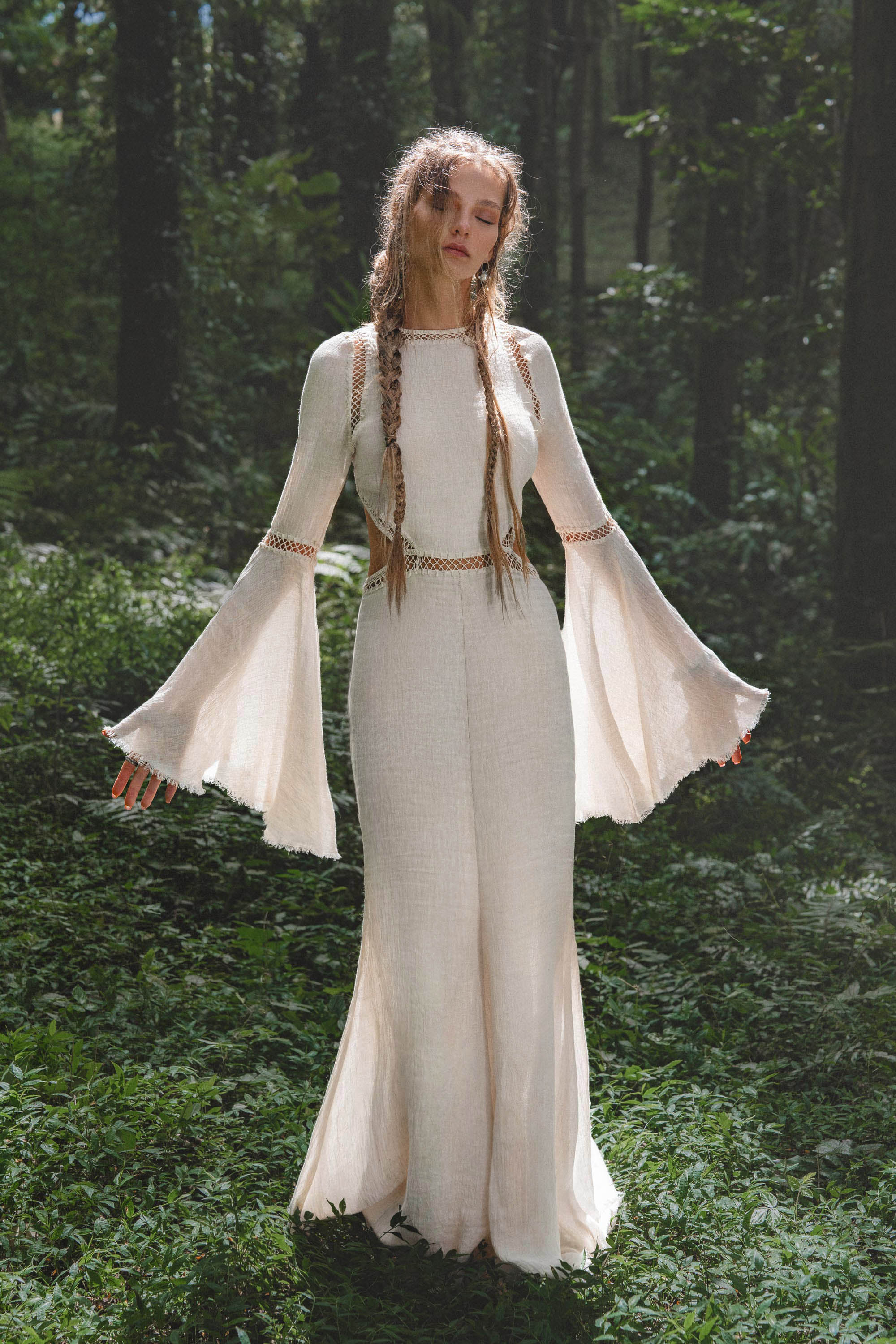 A woman with long, braided hair stands in a sunlit forest, eyes closed in serenity. She wears the AYA Sacred Wear Mandara Dress in Ivory – an organic boho wedding dress with wide bell sleeves and hand-stitched embroidery.