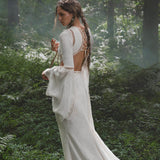 A woman with long, wavy brown hair stands among ferns in dappled sunlight, wearing the Mandara Dress in Ivory by AYA Sacred Wear—an organic boho wedding dress crafted from linen crinkle with hand-stitched embroidery.