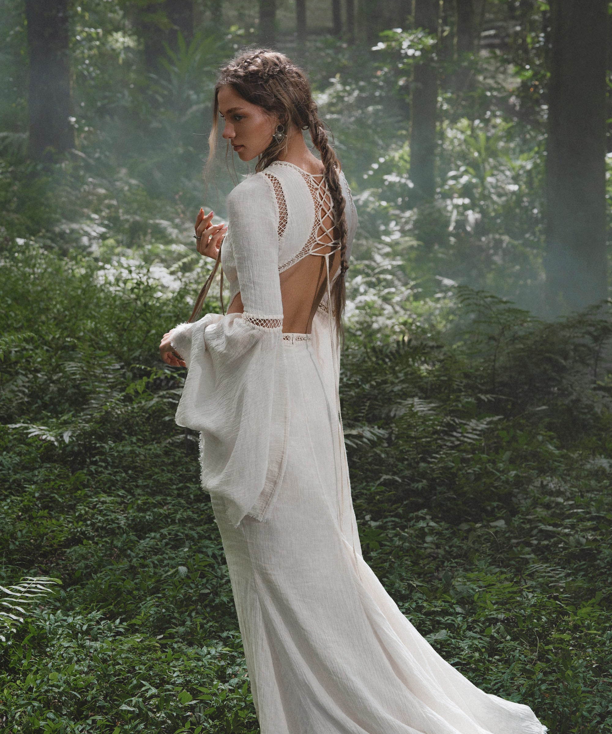 A woman with long, wavy brown hair stands among ferns in dappled sunlight, wearing the Mandara Dress in Ivory by AYA Sacred Wear—an organic boho wedding dress crafted from linen crinkle with hand-stitched embroidery.