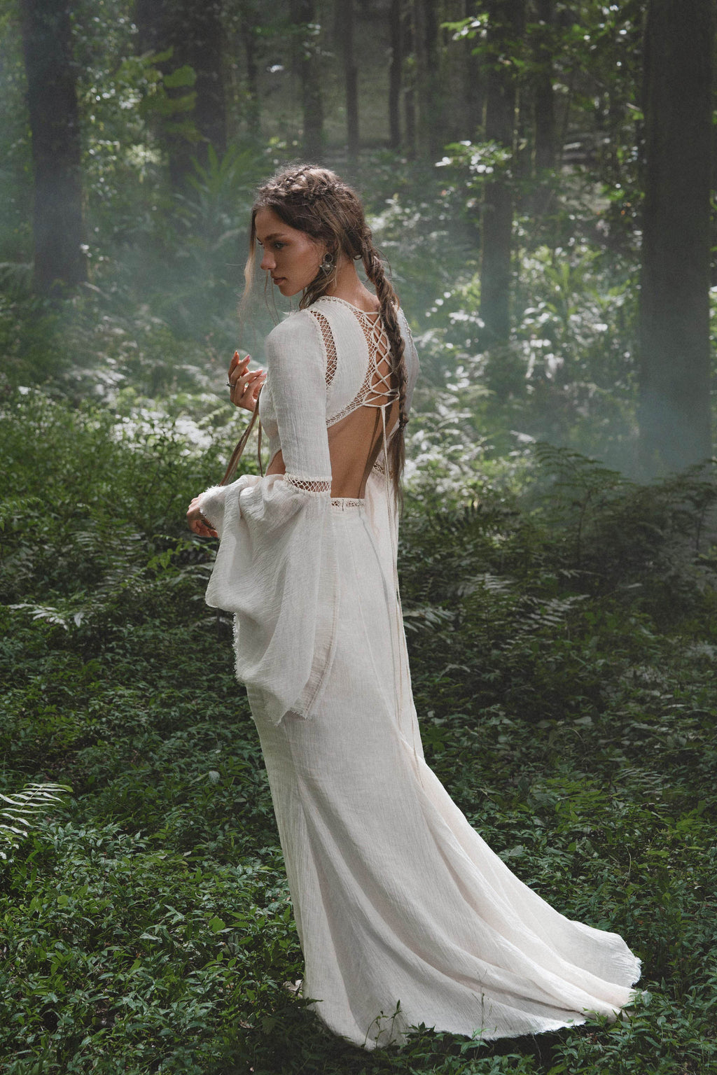 A woman with long, wavy brown hair stands among ferns in dappled sunlight, wearing the Mandara Dress in Ivory by AYA Sacred Wear—an organic boho wedding dress crafted from linen crinkle with hand-stitched embroidery.