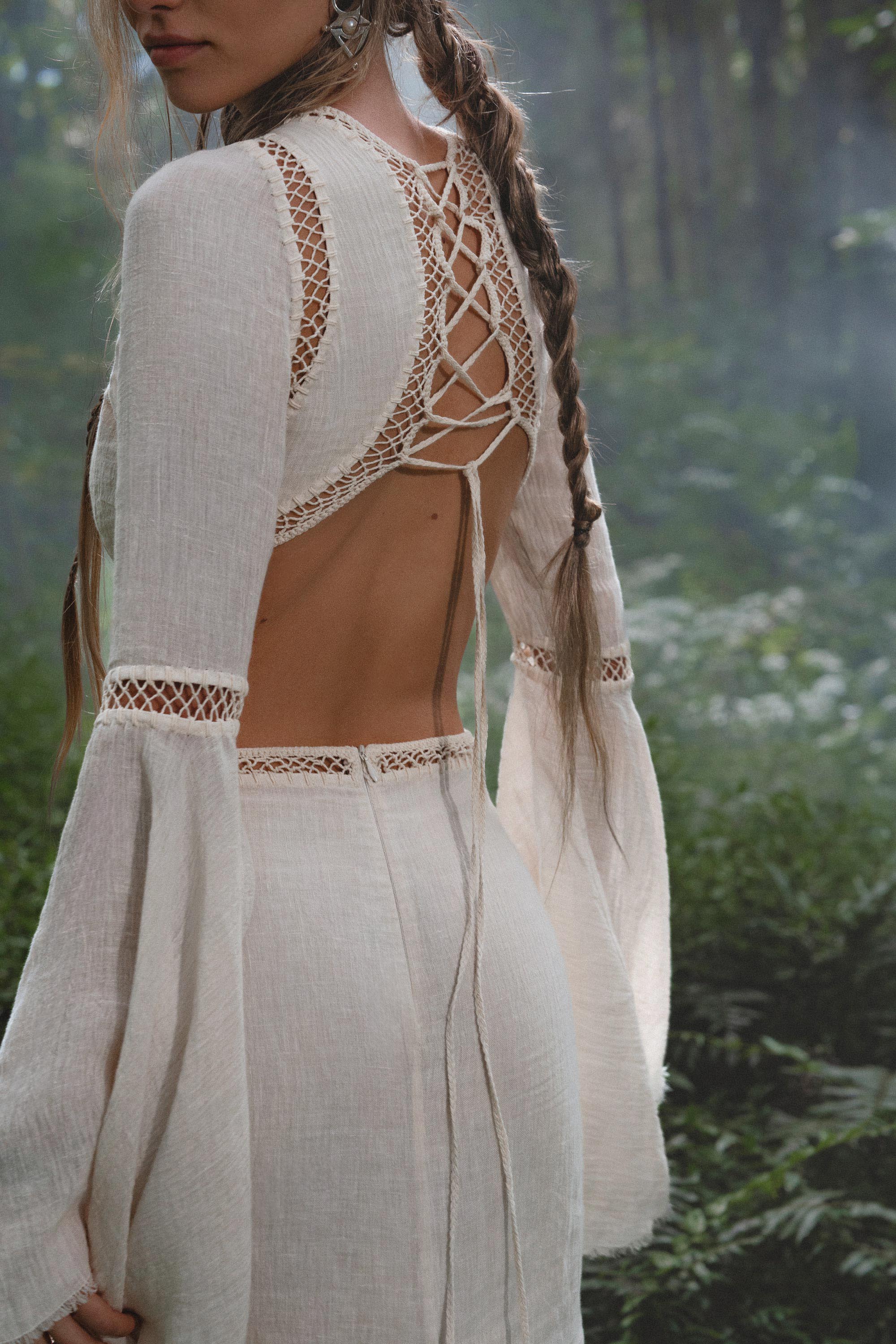In a misty green forest, a woman wears the Mandara Dress in Ivory by AYA Sacred Wear—an organic boho wedding dress with openwork lace, crisscross lace-up back, linen crinkle fabric, and hand-stitched embroidery for a dreamy, ethereal look.